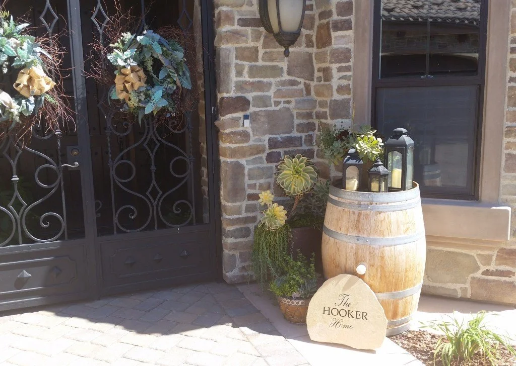 Decorative outdoor corner with a wooden barrel holding candles and lanterns, potted succulents and plants, stone and brick wall, decorative metal gate with hanging floral wreaths, and a stone sign reading 'The Hooker Home'.