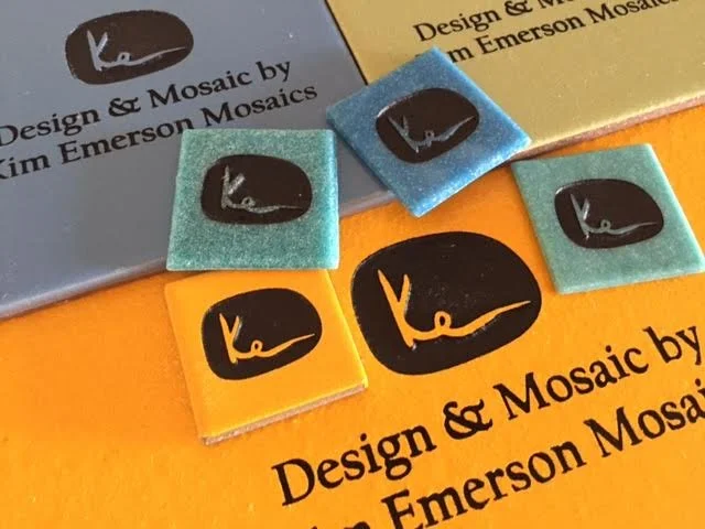 Colorful adhesive mosaic tiles with black and white logo, placed on papers about design and mosaics by Kim Emerson Mosaics.