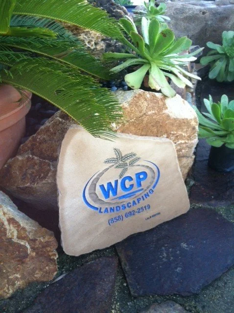 A stone with the logo of WCP Landscaping, surrounded by rocks and green plants.