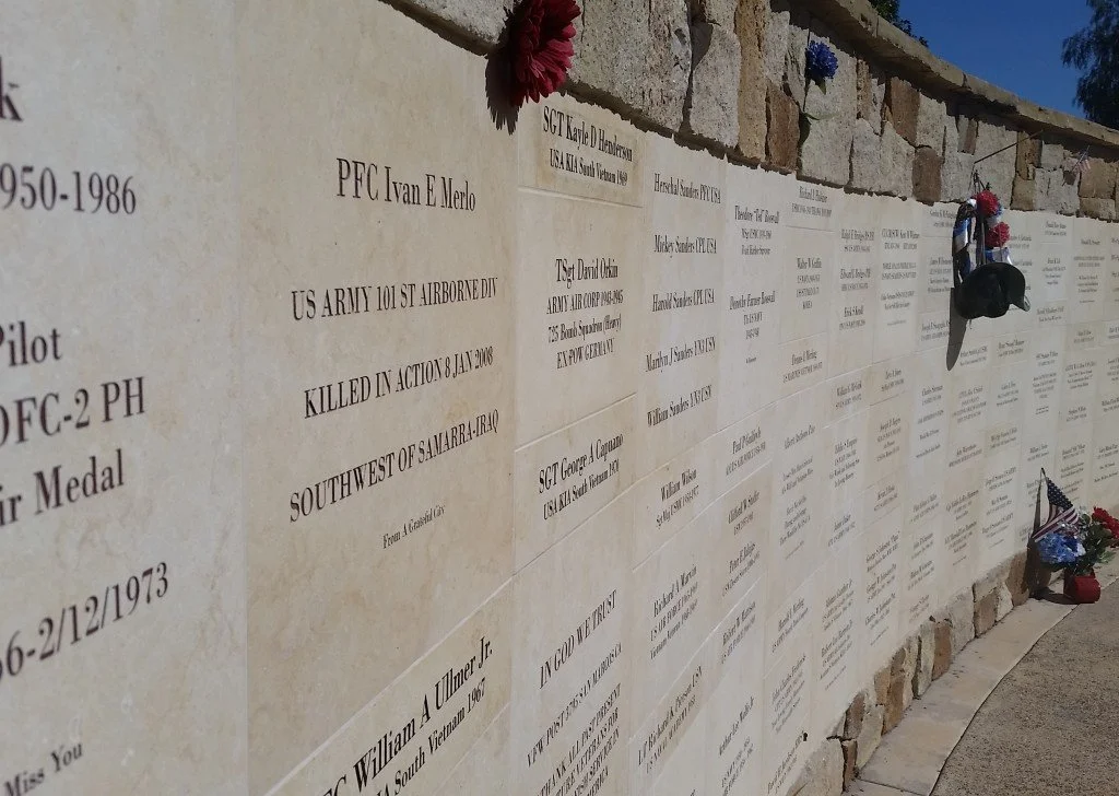 Memorial wall with engraved names and details of fallen soldiers, decorated with flowers, hats, and flags.