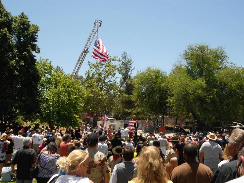 A large outdoor gathering with many people attending a patriotic event. An American flag is hanging from a tall fire truck ladder, and smaller flags are displayed on the stage and around the area. There are trees providing shade and a clear blue sky 