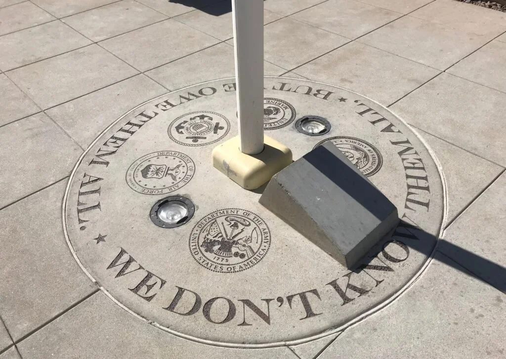 A circular plaque on the ground with the seal of the United States Department of the Army, surrounded by the words "ALL GAVE SOME, SOME GAVE ALL, BUT WE DON'T KILL". A pole with a base is attached to the plaque, and there are three small lights or sp