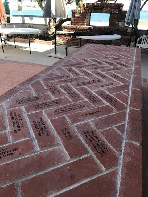 Sculpted brick wall with engraved plaques in an outdoor setting with tables and chairs, overlooking a water view.