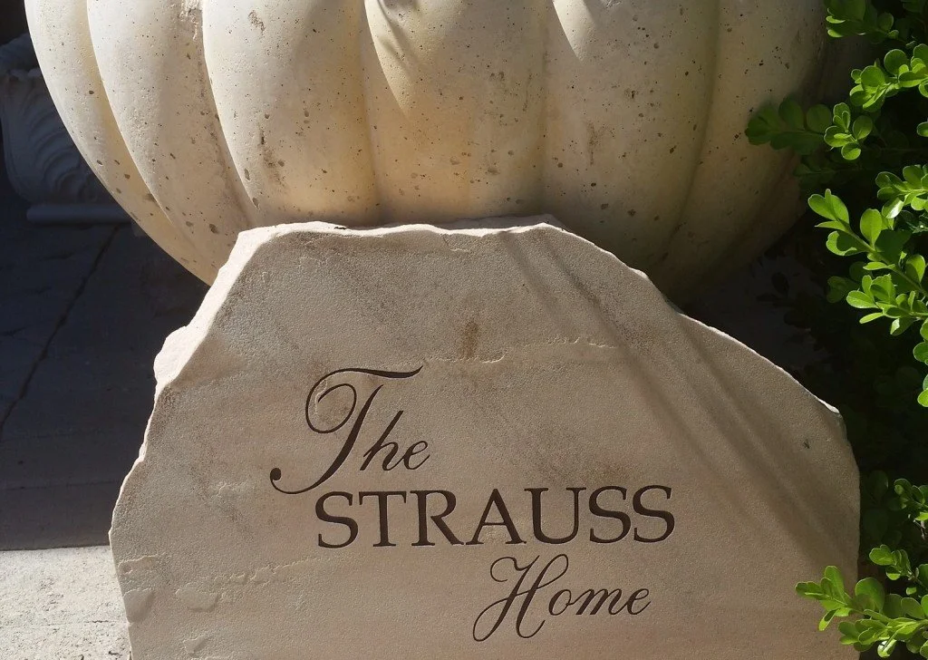 A white stone with the inscription 'The STRAUSS Home' in black lettering, placed outdoors near a large cream-colored pumpkin and green foliage.