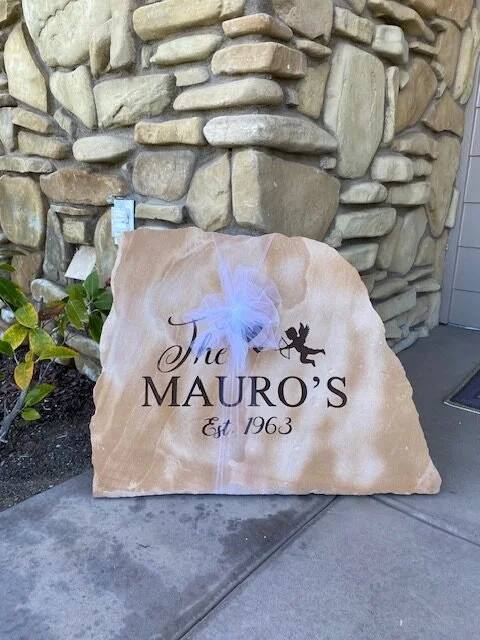 A beige stone sign with engraved black text reading 'The Mauro's Est. 1963,' decorated with a white tulle bow and a lizard silhouette, placed outside a house against a stone wall