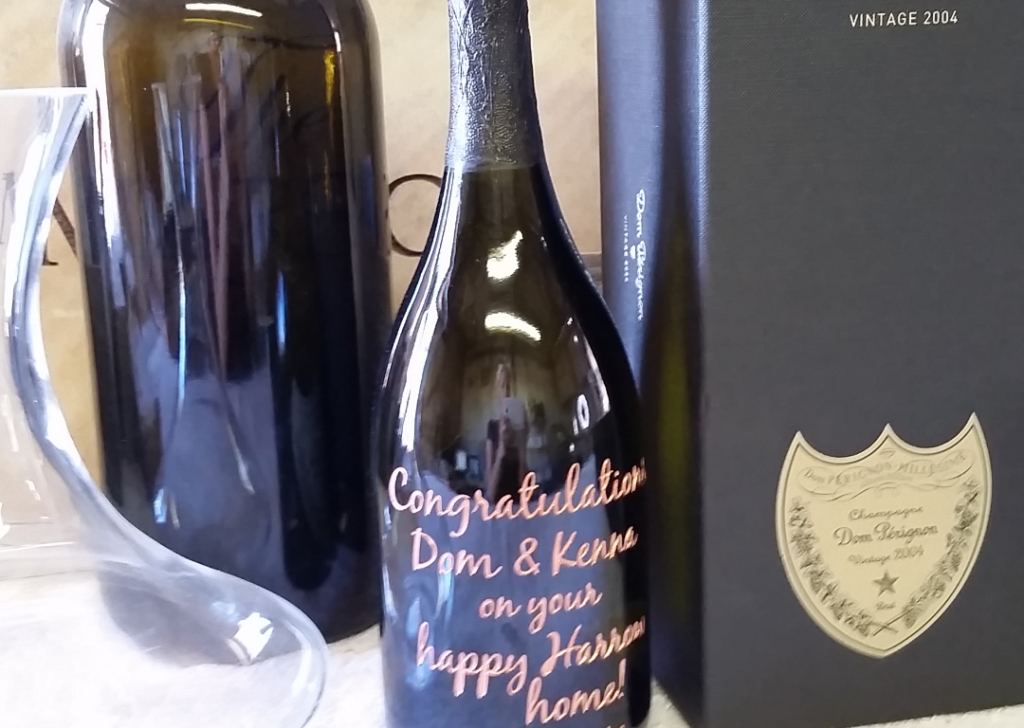 A champagne bottle with a personalized message in pink letters reading "Congratulations Dom & Kenna on your happy Hauser home!" next to a large black box and a trophy, with other bottles and glassware in the background.