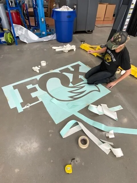 Child sitting on the floor working on a large decal with a logo that has a flame inside a circle, surrounded by the word 'FIRE'. The workspace includes tools, rolls of decals, and a blue trash can.