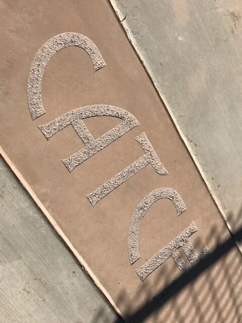 A brass or metal tile with the word "CATCH" engraved or etched into it, placed on a concrete sidewalk.