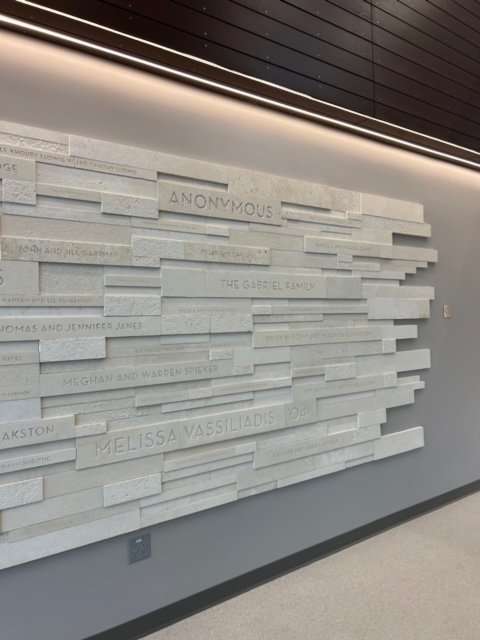 White brick wall with engraved names and the word 'ANONYMOUS' in the center, inside a modern building with a gray wall and ceiling lights.