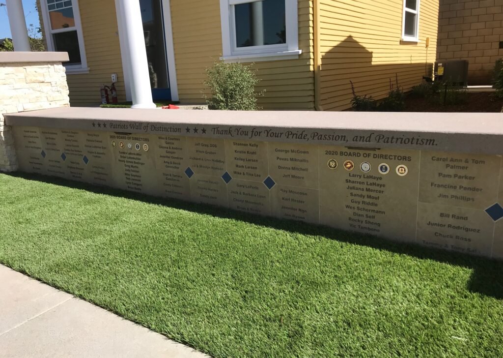 A stone and concrete memorial wall with engraved text and symbols, dedicating thanks to individuals and directors, located outdoors in front of a yellow house with green grass.