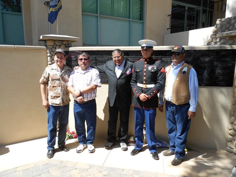 Five men standing in front of a memorial wall with plaques, outdoors in sunny weather. The men are dressed in various casual, military, and formal attire.