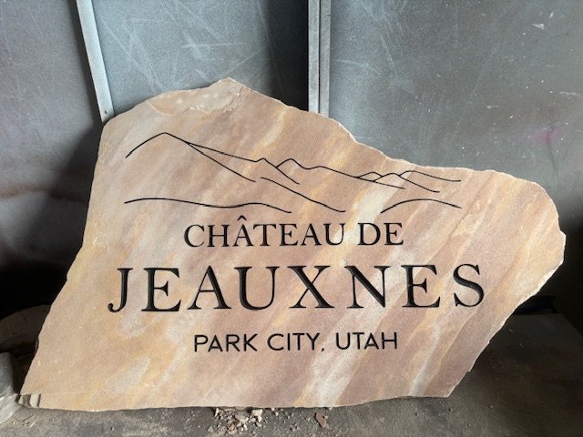 A large, irregularly shaped stone with engraved text reading 'Château de Jeauxnes, Park City, Utah' and a simple mountain line illustration, placed outdoors on a concrete surface