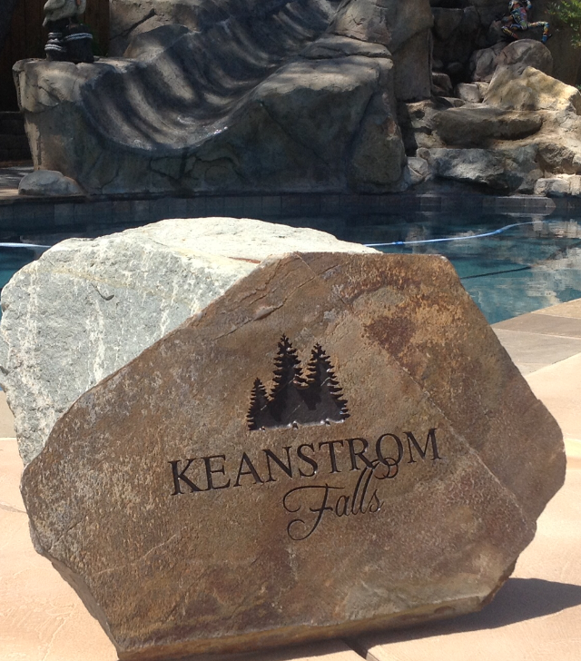 A large brown stone with engraved text and trees, located near a pool at Keanstrom Falls. In the background, there is water, rocks, and a partially visible person sitting on a rock.