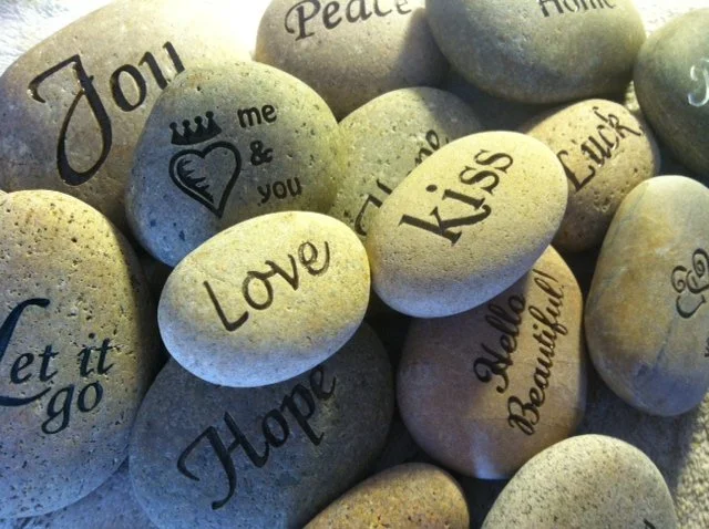 Collection of smooth, light-colored stones with inspirational and affectionate words and phrases written on them, such as "Love," "Hope," "Kiss," and "Jou".