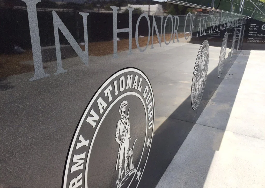 Reflection of a glass plaque displaying the National Guard emblem and part of a message that reads 'IN HONOR OF THE'.