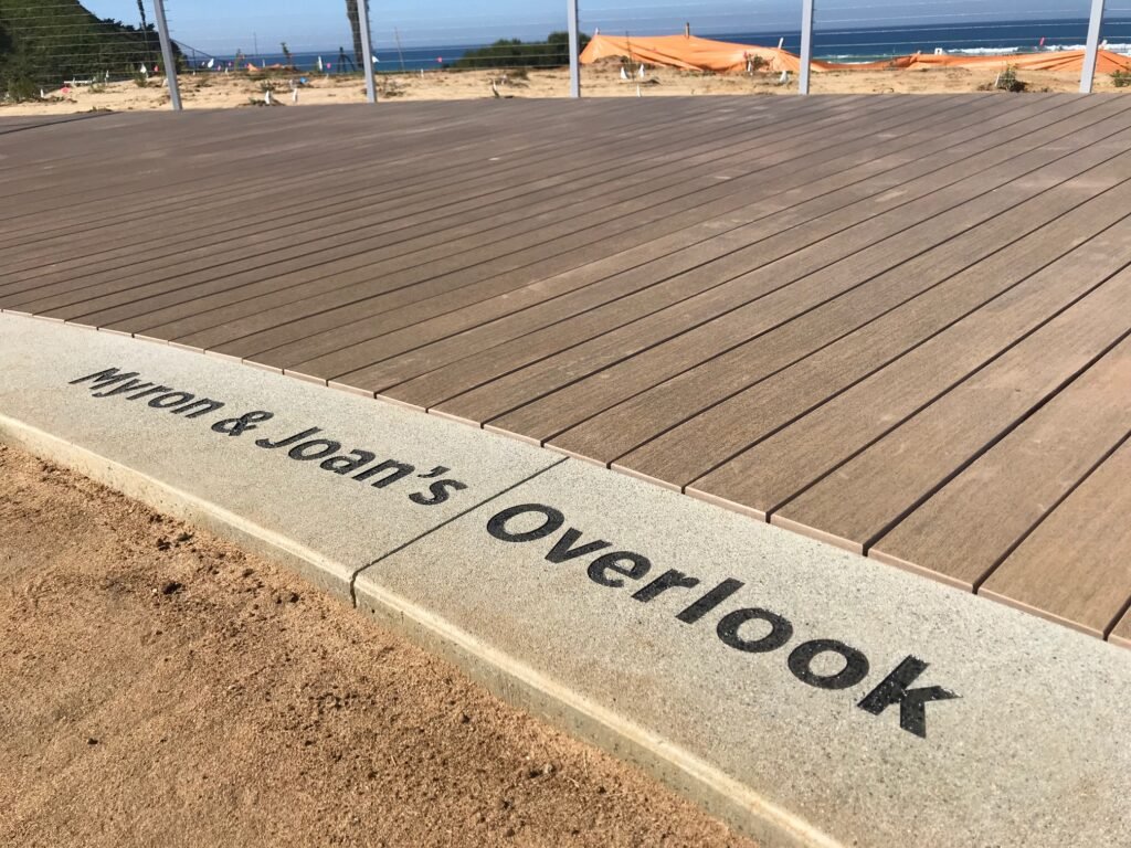 Close-up of a concrete curb with the words 'MYO & Joan's Overlook' painted on it, adjacent to a wooden deck overlooking a coastal area with a fence, some orange construction barriers, and the ocean in the background.