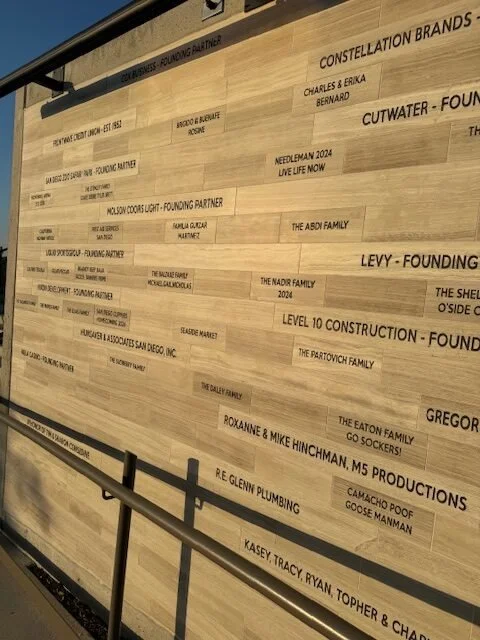 A wooden wall with engraved text listing names of companies or individuals, presumably in a sponsorship or partnership display. The wall has multiple horizontal lines of text with various company or family names.