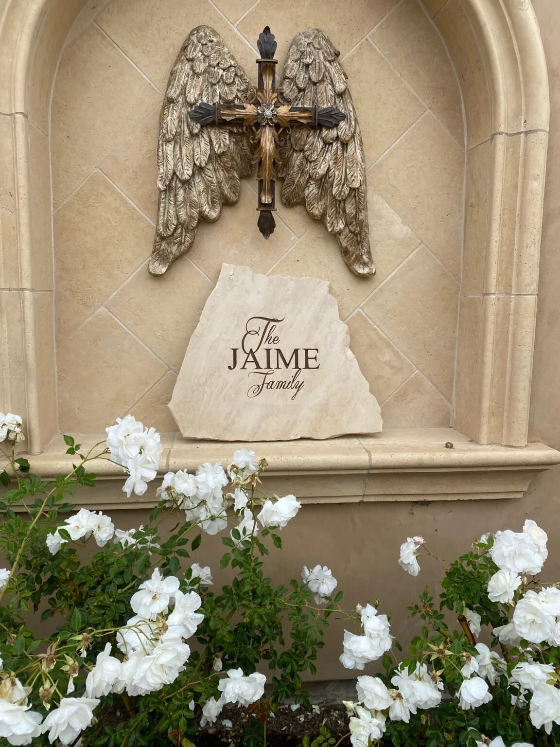 A stone memorial with a carved angel wing sculpture and a cross, inscribed with 'The Jaime Family,' surrounded by white flowers.