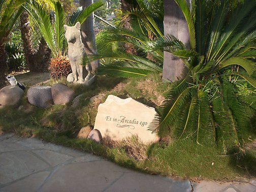 A garden scene with a stone statue of a lion, a large engraved stone that says "Et in Arcadia ego," green bushes, and palm trees.