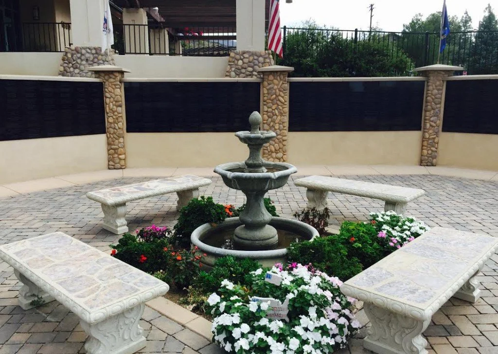 A small courtyard with a fountain in the center, surrounded by four stone benches and colorful flowers, enclosed by a wall with stone accents.