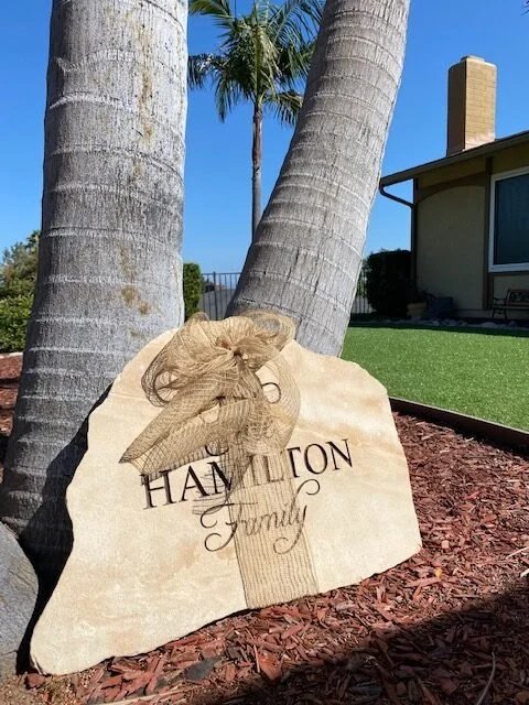 A decorative stone with the text 'Hamilton Family' and a gold bow in front of two palm trees and a house with a chimney, under a blue sky.