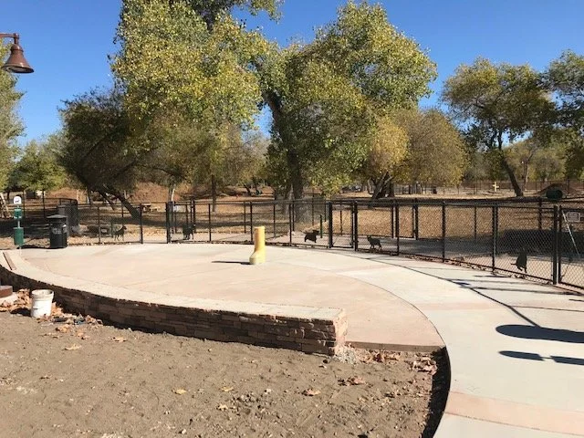 A paved outdoor area with a curved sidewalk, surrounded by a black metal fence, trees in the background, and a park setting.