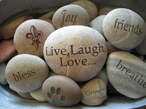 Collection of smooth decorative stones with engraved words and symbols, including 'Live, Laugh, Love...', 'joy,' 'friends,' 'bless,' 'grow,' a paw print, and 'breathe.'