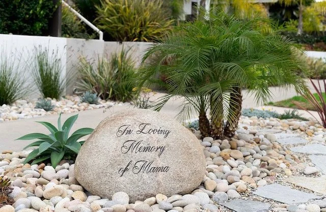 A memorial stone engraved with 'In Loving Memory of Mama,' located outdoors among decorative rocks and plants.