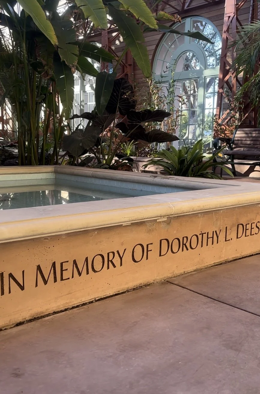 A memorial inscription on a stone ledge in a botanical garden or conservatory, with lush tropical plants and a large arched window in the background.