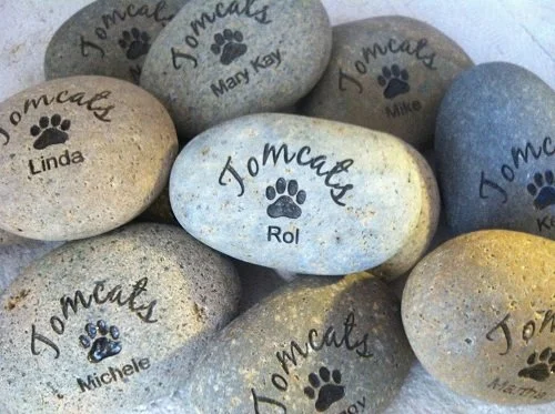 Smooth, oval-shaped stones engraved with the name 'Tommcays' and a paw print, each labeled with a different pet's name including Rol, Linda, Michelle, M Kay, Mike, and K K, arranged on a flat surface.