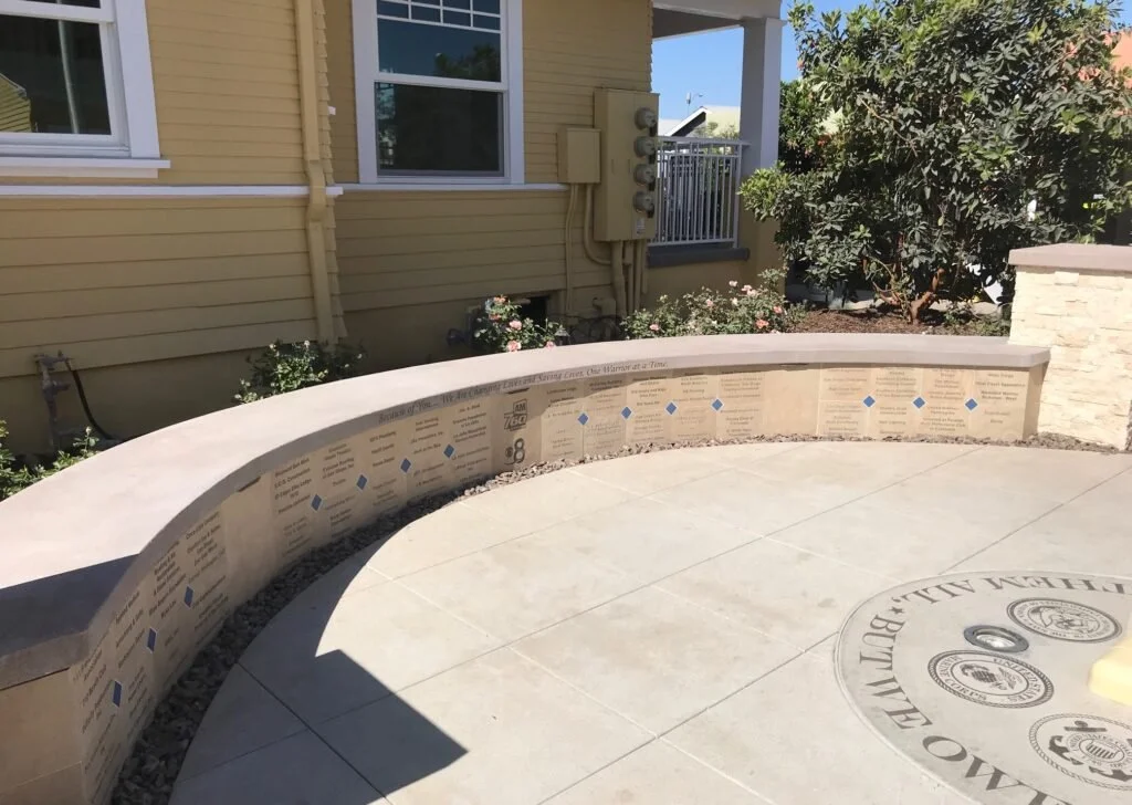 A curved concrete memorial wall with engraved text and blue accents, outdoor setting with houses, green bushes, and flowers in the background.