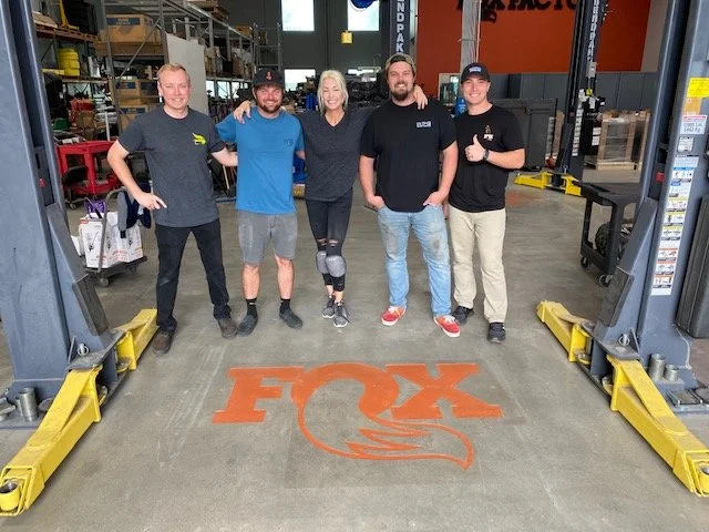 Group of five people standing inside a warehouse or industrial facility, in front of a painted fox logo on the floor, with equipment and shelving in the background. All are smiling and posing for the photo.