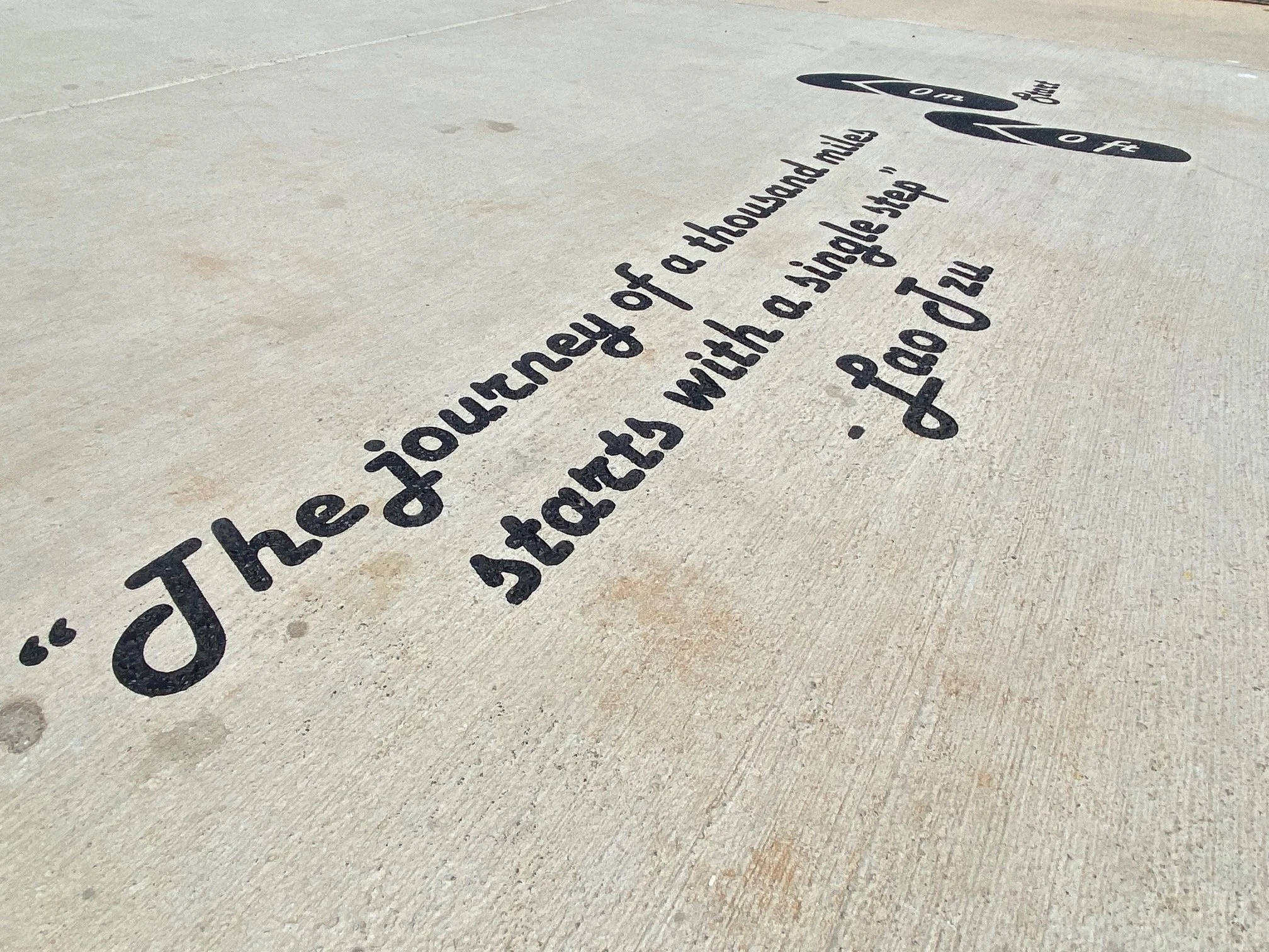 Close-up of a concrete floor with black stencil text stating, "The shoulder a touched and held and cried with a rattle when I was asleep" along with a black silhouette of a running figure.