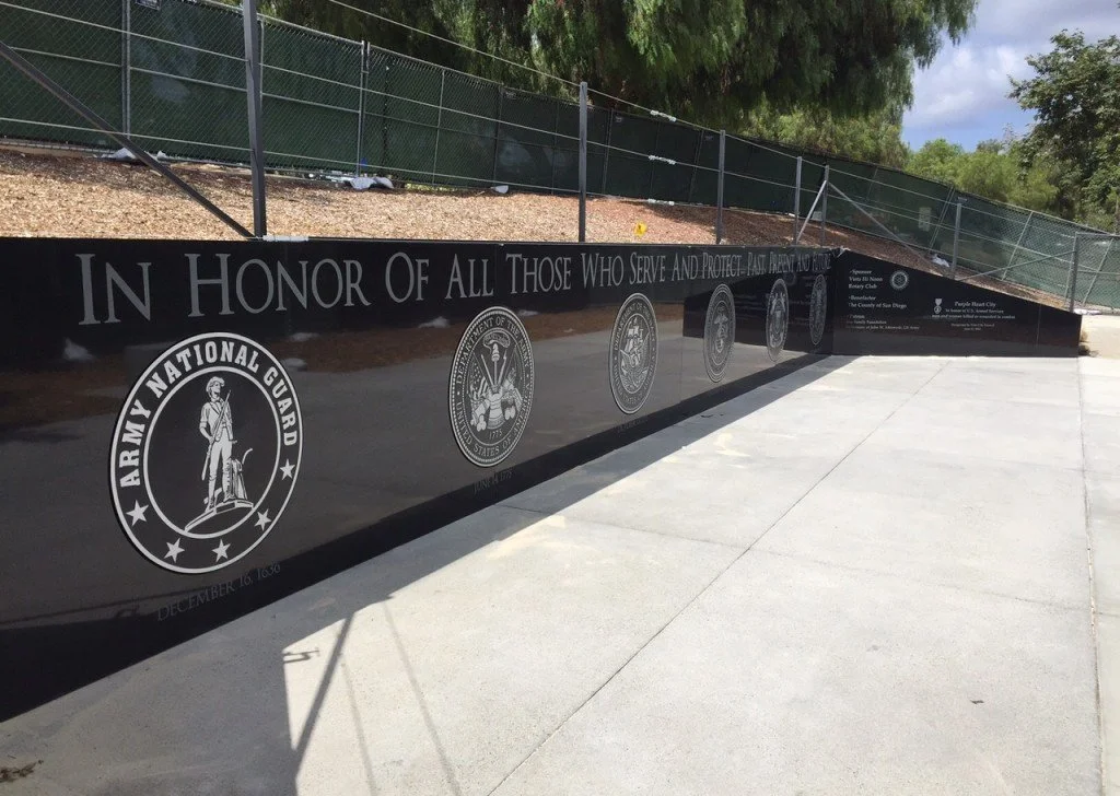 Black memorial wall with military seals and the inscription 'In Honor of All Those Who Serve and Protect Past Present and Future' on a sidewalk, with trees and a fence in the background.