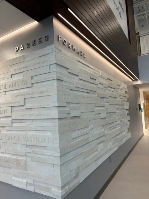 Interior view of a modern building with a textured white brick wall that has names and words engraved, featuring words like 'PARKER' and 'FORWARD' on the upper part, and the wall extends into a corridor with a gray floor.
