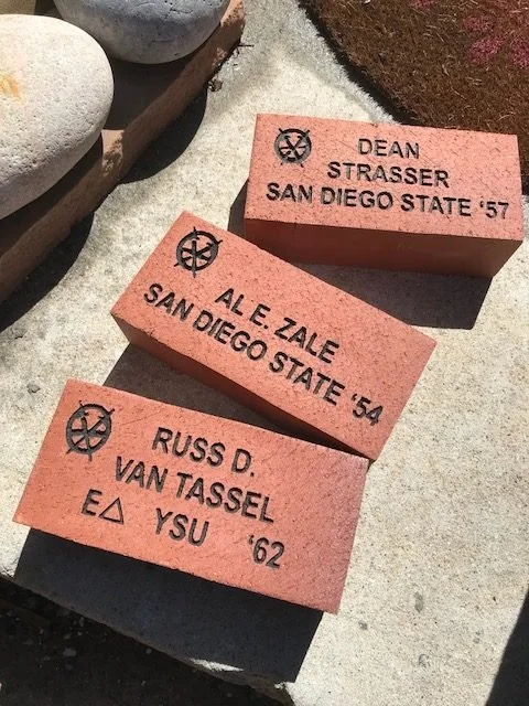 Three pink bricks with engraved names and years, placed on a concrete surface next to decorative stones. The bricks are inscribed with names of people and their affiliated institutions and graduation years.