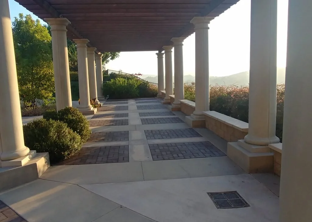 A covered outdoor walkway with beige columns, bricks and concrete paving, and surrounding greenery, overlooking a scenic view of distant hills during sunset.