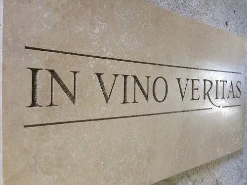 A beige-colored rectangular tile with the Latin phrase 'IN VINO VERITAS' engraved on it, tilted at an angle.