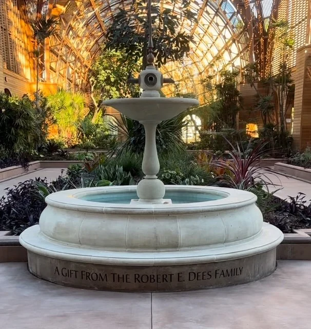 Indoor garden with a white tiered fountain and lush greenery, enclosed in a glass structure with sunlight streaming through.