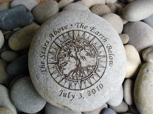 A gray stone engraved with a tree and circular clock design, surrounded by pebbles, with the inscription 'The Stars Above The Earth Below' and the date July 3, 2010.