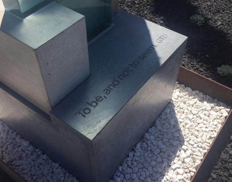 Close-up of a concrete bench with the engraved message "Table and chair of seed GTD," surrounded by white gravel and a planter with dark soil and small plants.