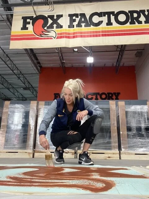 Woman painting a large red and white fox logo on the floor at Fox Factory store, with a Fox Factory sign overhead.