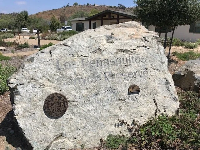 Large rock with engraved text and two patches, one with a seal and the other with a logo, in front of a park or community area with trees, a building, and vehicles in the background.