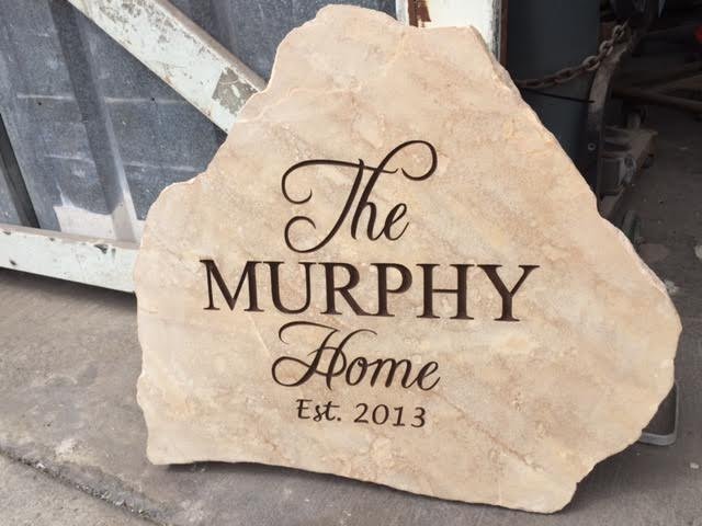 Stone sign with engraved text reading 'The Murphy Home Est. 2013' leaning against a metal structure in an industrial setting.