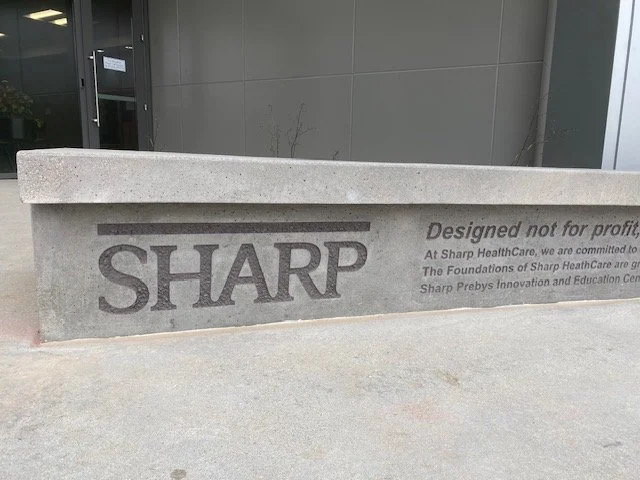 Concrete bench with the Sharp logo and some text engraved on it, placed outdoors near a gray building.