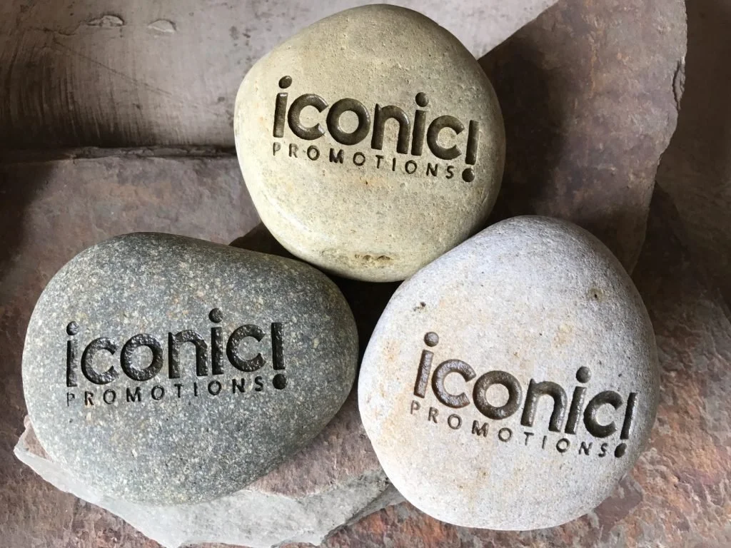 Three rocks with engraved text that reads 'iconic PROMOTIONS!' are placed on a stone surface.