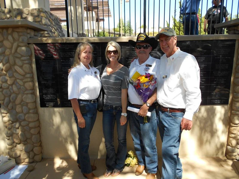 Four people standing in front of a memorial wall, with some holding flowers. The group includes three men and one woman, some wearing sunglasses and casual clothing, outside under a bright sky.