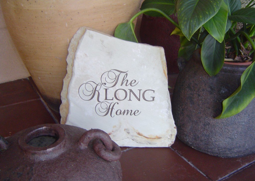 A decorative stone plaque with the words "The Long Home" written on it, placed among potted plants with large green leaves.