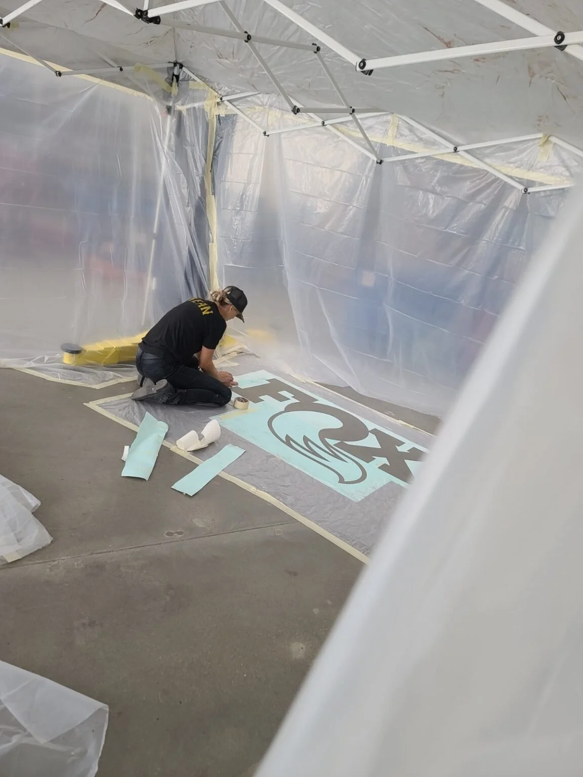 A person in black clothing and a cap kneels on the floor, working on a large banner with a design that includes a whale and the word "RESCUE". The area is temporary covered with plastic sheets.
