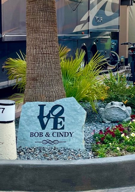 A large gray rock with the word 'LOVE' carved into it, along with the names 'BOB & CINDY.' A small frog sculpture, green plants, and a palm tree are nearby, with a bus and other bicycles in the background.